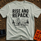 Rise And Repack - T-Shirt in Bay - A black-and-white illustration of a man kneeling to roll up a sleeping bag, with a...